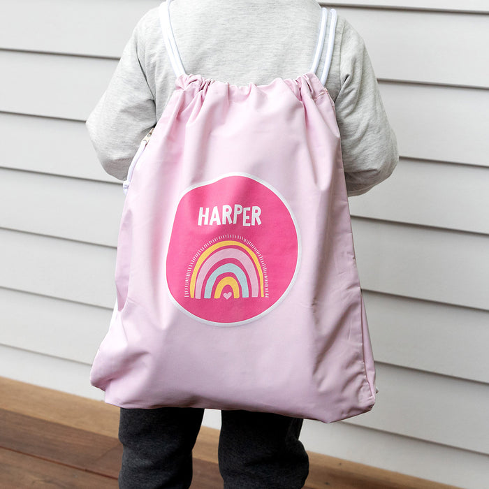 Child wearing pink drawstring swim bag with rainbow print and personalised name.
